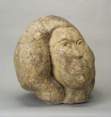 Rzeźba przypominająca owalny, beżowy kamień, jej prawa część to twarz z uproszczonymi rysami twarzy i szyja, lewa to ukazane za pośrednictwem jednej obłej formy włosy.