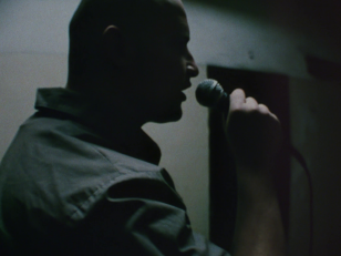 Kadr z filmu utrzymany w zimnej, niebieskawej tonacji, przedstawia profil mężczyzny widocznego od ramion w górę i trzymającego w dłoni mikrofon.