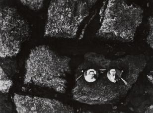 Czarno-biała fotografia w układzie poziomym. Kadr wypełniają leżące obok siebie kamienie. W prawej dolnej części na jednym z kamieni leżą okulary. W szkła okularów wmontowane są fotografie z wizerunkami kobiety i mężczyzny.