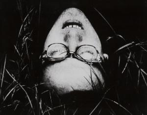 Czarno-biała fotografia w układzie poziomym. Zdjęcie przedstawia twarz mężczyzny w okularach. Mężczyzna leży w trawie, ma rozchylone usta, zamknięte oczy.