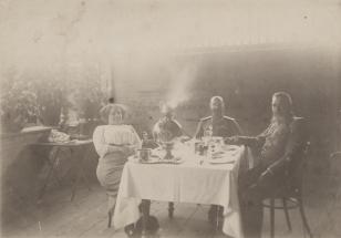 Fotografia czarno-biała przedstawia trzy osoby siedzące przy stole przykrytym obrusem, z samowarem po środku. Od lewej siedzi odchylona lekko do tyłu kobieta ubrana w jasną bluzkę i spódnicę, dalej dwóch mężczyzn w mundurach.