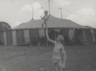 Fotografia przedstawia młodego mężczyznę ubranego w slipy przed namiotem cyrkowym trzymającego na wyprostowanej ręce nagą, około trzyletnią dziewczynkę.