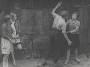 Fotografia przedstawia dwie dziewczynki z prawej strony przyglądające się chłopcu i dziewczynce z lewej strony. Chłopiec stoi odwrócony tyłem, oparty ręką o drewnianą scianę, i wyciąga drugą rękę w kierunku dziewczynki zaczepiając ją. Ona usiłuje go powst