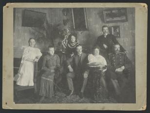 Fotografia przedstawia portret rodziny w mieszkaniu. Cztery osoby siedzą, trzy stoją za nimi.  Postacie ubrane są elegancko, za nimi wiszą obrazy, we wnętrzu znajdują się kwiaty.