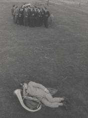 Fotografia przedstawia leżącego na pierwszym planie mężczyznę z puzonem i grupę 16 mężczyzn z instrumentami. Ubrani są w szare dwurzędowe garnitury i czapki strażackie. Obok nich lekko z tyłu jaśniej ubrany mały chłopiec.
