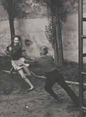 Fotografia przedstawia scenę, w której chłopak goni dziewczynę. Zza dziewczyny wystaje ręka i noga trzeciego dziecka. W tle widoczny mur i dwie topole. Z prawej strony zdjęcia metalowa drabina.