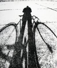 Czarno-biała fotografia, na pierwszym planie wyciągnięty cień roweru i rowerzysty. Obraz cienia tworzą nieregularne czarne plamy. Reszta niemalże biała.