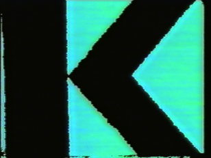 Kadr z filmu - czarna litera K wypełniająca cały kadr, umieszczona na turkusowo-niebieskim tle.