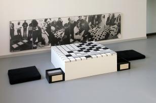Instalacja złożona z białego kubika, który stanowi planszę z białymi, szarymi i czarnymi kwadratowymi polami ułożonymi jak na szachownicy,  po bokach dwa pudełka  i dwie czarne poduszki, w tle pięć czarno-białych fotografii dokumentujących przebieg gry z 