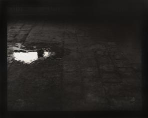 Fotografia czarno-biała, ciemna, niemal nieczytelna w cieniach, widziana z góry podłoga, z lewej u góry nieduża kałuża odbijająca jasne światło i okno.