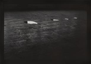 Fotografia czarno-biała, widziana z góry podłoga, na niej w nieco jaśniejszym miejscu cztery zmniejszające się kałuże, rzędem w wodzie odbijają się fragmenty pomieszczenia.