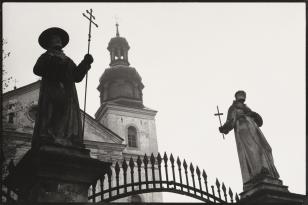 Fotografia czarno-biała, kadr poziomy. Na pierwszym planie przy dwóch bocznych krawędziach zdjęcia stojące na cokołach dwie figury świętych. Trzymają w rękach krzyże. Między nimi ostro zakończone przęsła bramy. W tle wieża kościoła oraz szare niebo.