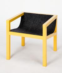 Żółty fotel w kształcie sześcianu z siedzeniem pokrytym ciemnym filcem, wsparty na czterech graniastych nogach łączących się z poręczami. Siedzenie jako jedyne zamiast kąta prostego ma postać płynnej linii.