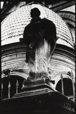 Fotografia czarno-biała, kadr pionowy. Zbliżenie na posąg świętego. Podest i górna część postaci ciemniejsza nisz pozostała część kompozycji. W tle kopuła z pokryciem dachowym w formie łusek.
