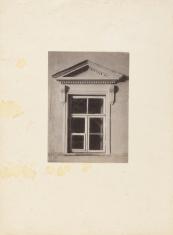 Fotografia architektoniczna w sepii, kadr pionowy. Zbliżenie na otynkowaną ścianę z oknem oraz zdobieniem nad nim w postaci portyku.