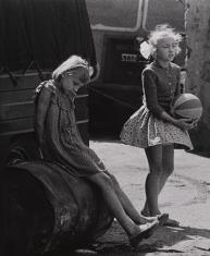Fotografia czarno-biała przedstawiająca dwie dziewczynki na podwórku, dziewczynka z lewej trzyma piłkę, z prawej siedzi na blaszanej beczce, w tle fragment samochodu.