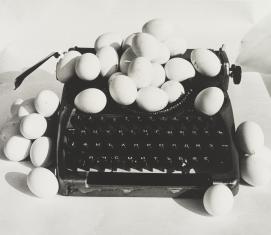Czarnobiała fotografia przedstawiająca stojącą na białym blacie maszynę do pisania, wkół której i na której znajdują się jajka.