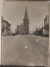 Czarno-biała fotografia w pionie, przedstawiająca perspektywiczne ujęcie brukowanej ulicy, na drugim planie kościół z wieżą otoczoną rusztowaniem, po bokach zabudowania miejskie, w centrum mężczyzna na rowerze, widać przechodniów i bryczki konne.