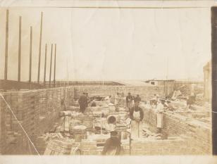 Fotografia w tonacji sepii, przedstawiająca scenę budowy domu - ubrani w jasne fartuchy i ciemne czapki mężczyźni układają cegły poszczególnych ścian, dookoła wiele drewnianych beczek.