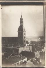 Fotografia czarno-biała w pionie, przedstawiająca widok dachów miasta, dominuje nad nimi wieża kościoła zwieńczona hełmem w centrum kompozycji.