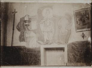 Czarno-biała fotografia pokazująca ścianę we wnętrzu kościoła - nad drzwiami fresk z postacią władcy, po lewej krzyż, po prawej obraz z cyklu Drogi Krzyżowej, na pierwszym planie wiszący metalowy przedmiot, lampka lub kadzielnica.