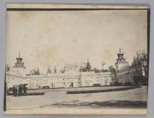 Fotografia (najprawdopodobniej w sepii lub wyblakła) ukazuje trójskrzydłowy jasny pałac, po lewej stronie trzy postacie z parasolkami i w ciemnych sukniach widoczne od tyłu.
