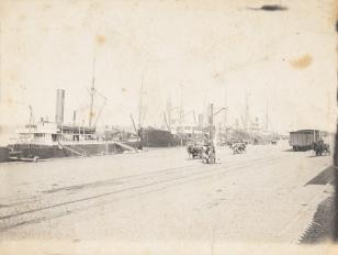 Fotografia czarno-biała, kompozycja w poziomie przedstawiająca widok doków z cumującymi statkami i biegnącą po ukosie drogą na której widać wozy konne i szyny z jednym metalowym wagonem po prawej stronie kompozycji.