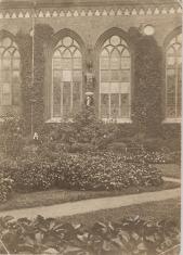 Fotografia czarno-biała. Kompozycja w pionie. Widok  fragmentu elewacji kościoła z oknami gotyckimi. Przed nią widać skwer bogato porośnięty roślinnością.