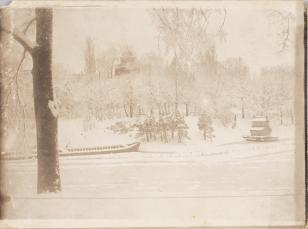 Fotografia czarno-biała w poziomie przedstawiająca pejzaż zimowy - pokryte śniegiem rzekę z porzuconą łodzią, drzewa, w tle wzgórze kopulastą wieżą kościoła, po prawej stronie niewielki budynek z podwójnym spadzistym dachem.