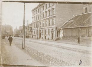 Fotografia czarno-biała przedstawiająca widok ulicy, lewą stronę kadru zajmuje chodnik, środkową brukowana jezdnia, a po prawej widoczne kamienice i drewniany parterowy dom. Po lewej stronie na pierwszym planie idzie ciemno ubrany mężczyzna, w tle więcej 