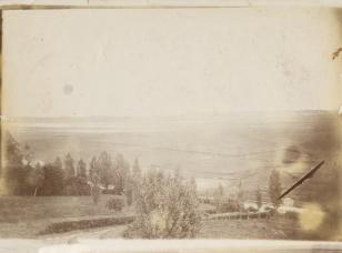 Fotografia czarno-biała. Kompozycja w poziomie przedstawiająca spokojny pejzaż z drzewami i panoramą łąk ciągnącą się ku horyzontowi.