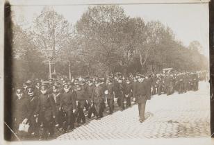 Fotografia czarno-biała, kompozycja w poziomie przedstawiająca mężczyzn i chłopców w mundurach, idących drogą wzdłuż drzew.