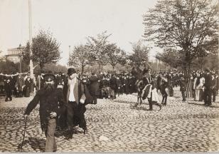 Fotografia czarno-biała. Kompozycja w poziomie. Scena miejska. widok licznej grupy mężczyzn w mundurkach gromadzących się brukowanej drodze. Po lewej widacdwóch starszych mezczyzn z brodami i w płaszczach - jeden w kaszkiecie, drugi w meloniku. Po prawej 