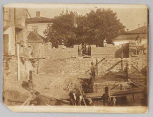 Fotografia w tonacji sepii w poziomie, przedstawiająca budowę domu i fundamentów przez grupę mężczyzn, dookoła okoliczne budynki..