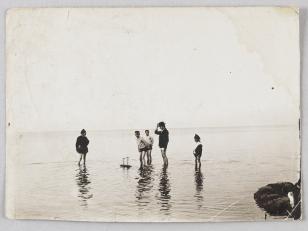Fotografia czarno-biała przedstawiająca scenę nadmorską. Ujęcie mężczyzny z czwórką chłopców stojących w wodzie.