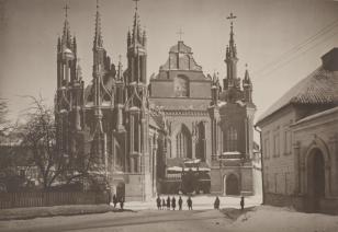 Fotografia czarno-biała  w poziomie. Fasada trójwieżowego kośćioła gotyckiego św. Anny i kościoła Bernadrynów. Ujęcie pora zimową. Drobne postaci ludzkie na pierwszym planie, a po prawej budynek z trzema zaślepionymi oknami na parterze i pierwszym piętrze