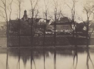 Odbitka fotograficzna - na pierwszym planie gładka tafla wody i szpaler siedmiu bezlistnych drzew; w tle wydłużony gmachu pośród zabudowań i wieża kościelna.