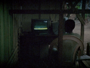 Kadr z filmu - pod wiatą, skleconą z płyt falistych, siedzi na białym, plastikowym, ogrodowym krześle osoba odwrócona tylem do widza, o ciemnej skórze i czarnych, kręconych, krótko ostrzyżonych włosach, przed nią ekran telewizora, na którym obraz jest nie