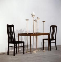 Instalacja złożona ze stołu i dwóch krzeseł bez siedzisk. Stół nie ma blatu, na drewnianych elementach unosi się nad nim dzbanek, filiżanki,  i spodeczki.