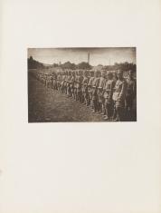 Fotografia wykonana w kolorystyce sepii, poziomy kadr. Ogólny widok na stojących w dwurzędowym szeregu żołnierzy. Ubrani są w pełne umundurowanie. Mężczyźni stoją na polu ze ściętą trawą. W tle za nimi widać kominy i drzewa.