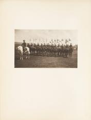 Fotografia wykonana w kolorystyce sepii, poziomy kadr. Żołnierze siedzący na ciemnych koniach z chorągwiami w dłoniach. Na przodzie z lewej strony obrazu siedzi jeden oficer na koniu wyróżniającym się jasną wręcz białą maścią. W tle pochmurne niebo.