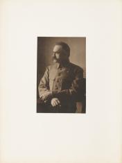 Fotografia portretowa w sepii, pionowy kadr. Marszałek Józef Piłsudski w mundurze. Mężczyzna jest sfotografowany z lewego profilu od linii bioder na siedząco. Światło pada na postać z lewej strony obrazu. Tło o jasnej kolorystyce, lekko doświetlone.