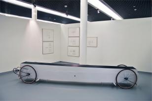 Pojazd o kształcie podłużnej skrzyni z 4 kołami rowerowymi. Blat pokryty czarną gumą. W tle przestrzeń wystawy.