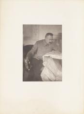 Fotografia wykonana w kolorystyce czarno-białej, kadr pionowy. Siedzący Marszałek Józef Piłsudski na krześle. Zwrócona jest ku przedmiotom leżącym na stole po prawej stronie obrazu; Mężczyzna ma założoną nogę na nogę.