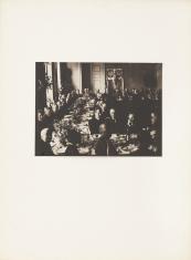 Fotografia wykonana w kolorystyce biało-czarnej, kadr poziomy. Mężczyźni ubrani na galowo siedzą przy stole ustawionym w literę U. Są w trakcie posiłku. W górnej części obrazu na ścianie widać godło polskie i litewskie.