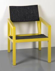 Żółty fotel z prostokątnym oparciem i siedzeniem pokrytym ciemnym filcem, wsparty na czterech graniastych nogach łączących się z poręczami.