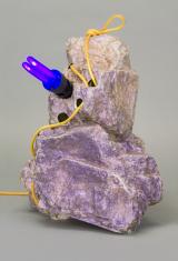 Zdjęcie kształtu z tworzywa sztucznego przypominającego skałę o jasnofioletowej tonacji, przez otwory w niej przechodzi żółty kabel, prowadzący do niebieskofioletowej żarówki w czarnej oprawie zamocowanej w górnej części formy.