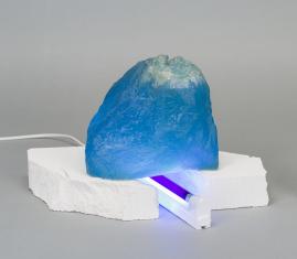 Zdjęcie rzeźby złożonej z trzech elementów: niebieskiej transparentnej formy przypominającej górę, jej białej podstawy z betonu i przechodzącej przez jej środek fioletowej świetlówki w białej oprawie zamocowanej w podstawie.