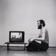 Czarnobiałe zdjęcie przedstawia rozebranego od pasa w górę mężczyznę siedzącego ze skrzyżowanymi nogami profilem do widza, zwrócony jest twarzą do telewizora na niskim stołeczku, na ekranie powtarza się ten właśnie obraz, sekwencja trwa w nieskończoność.