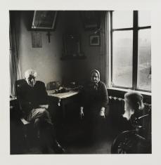 Czarno-biała fotografia portretowa. Przedstawia parę starszych osób we wnętrzu mieszkania. Mężczyzna i kobieta siedzą przy stole w rogu pomieszczenia. Na obu przyległych ścianach są okna, które oświetlają izbę. W rogu na pierwszym planie widać wrzeciono.